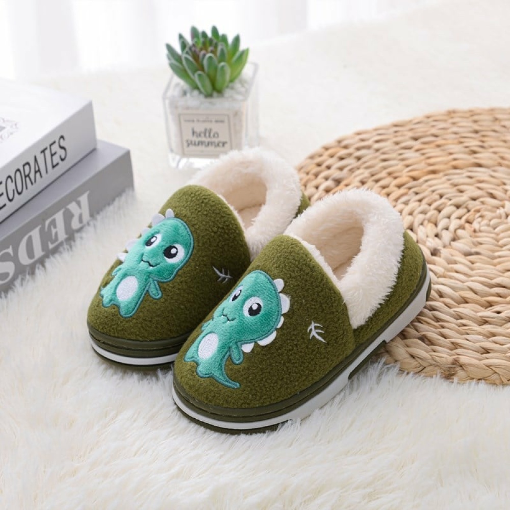 Pantofole di cotone verde dinossaure per bambini, davanti a una pianta su un tappeto bianco