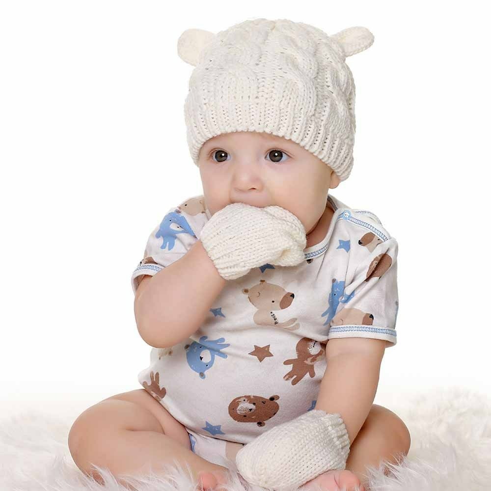 Set di cuffiette e guantini invernali bianchi per neonato con il bambino che tiene la mano in bocca e abbigliamento a fantasia bianca
