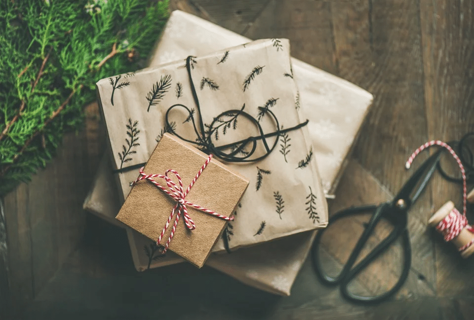 3 pacchi regalo grigi e marroni, circondati da nastri regalo, posizionati su un pavimento di legno marrone con uno scalpello e un ramo d'albero sullo sfondo