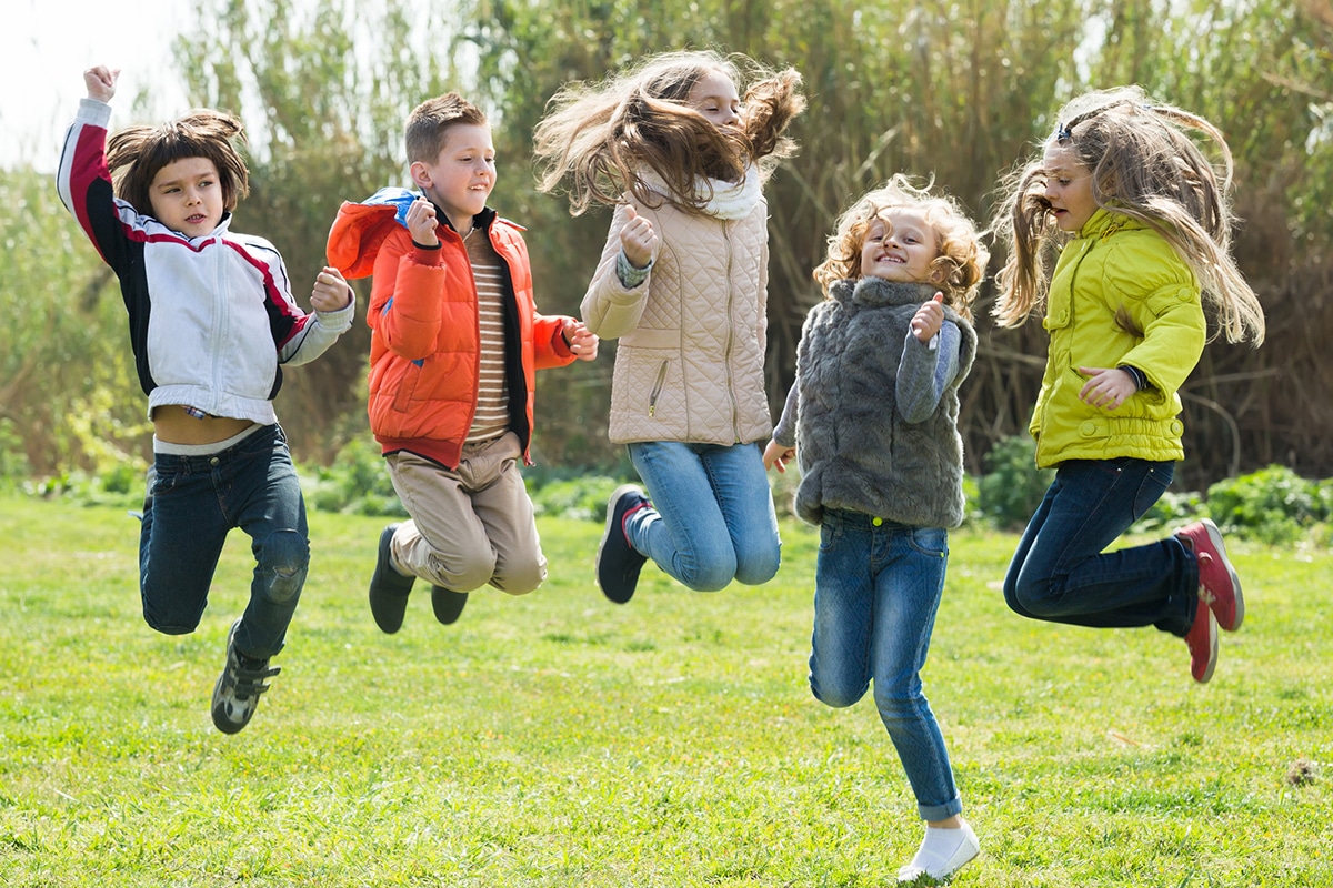 Un gruppo di bambini felici che saltano sull'erba all'aria aperta. Ci sono 2 bambini e 3 bambine. Ci sono degli alberi sullo sfondo