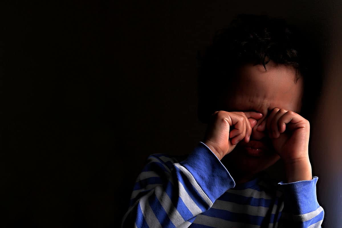 Un bambino di razza mista con un maglione a righe bianche e blu. Il bambino si tiene gli occhi perché ha paura del buio.