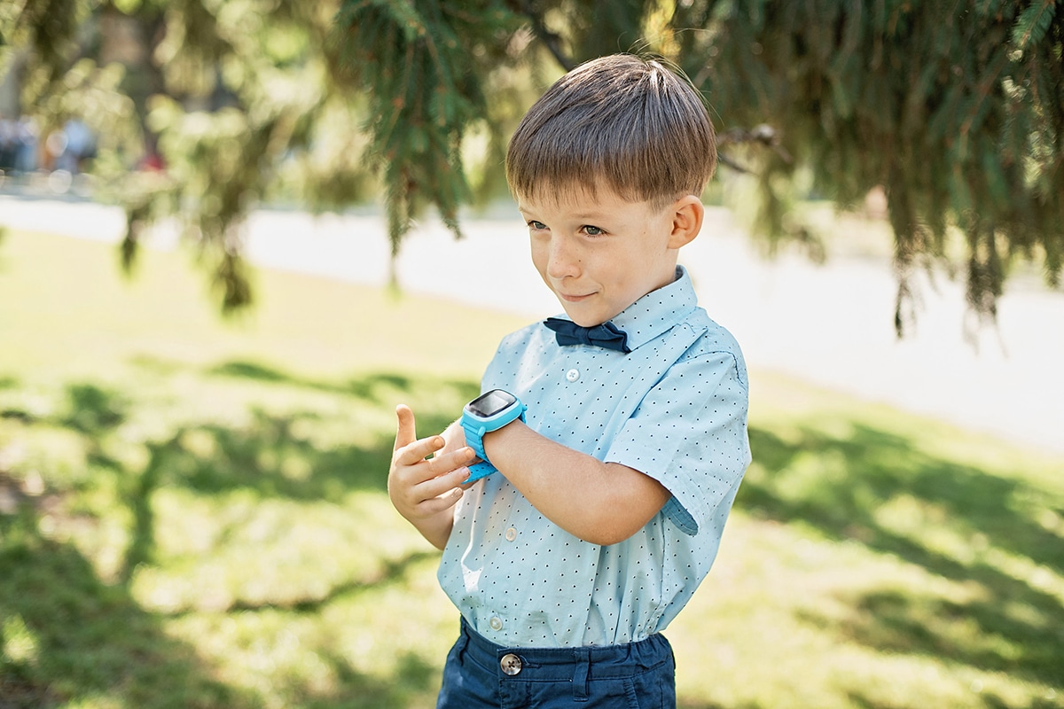 Un ragazzo che parla al suo orologio connesso. Indossa un orologio blu, una camicia blu e si trova all'aperto sotto il sole. Ci sono degli alberi sullo sfondo