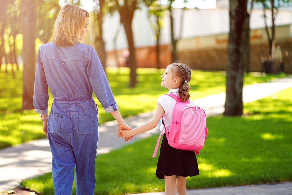 Una madre porta sua figlia in un parco. C'è il sole, la madre è vestita in jeans blu, la bambina indossa uno zaino rosa per bambini
