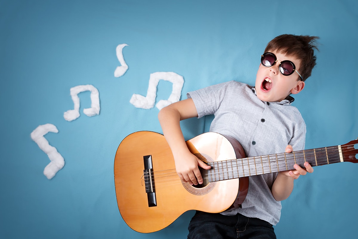 Un ragazzo che suona la chitarra su uno sfondo blu. Il ragazzo indossa occhiali scuri, una camicia grigia a maniche corte e una chitarra di legno marrone. Ci sono note musicali bianche sullo sfondo