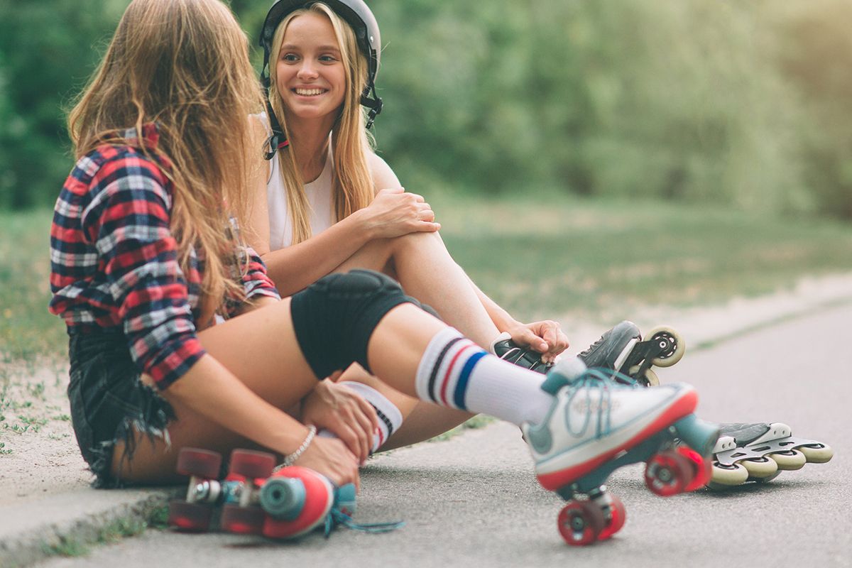 Due giovani ragazze sedute sul ciglio della strada. Una indossa pattini a rotelle, l'altra pattini a rotelle e casco.