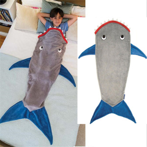 La foto è divisa in due parti: la prima mostra un bambino sdraiato a letto, infilato in un sacco a pelo grigio a forma di squalo con pinne blu e bocca rossa. La seconda parte dell'immagine mostra questo sacco a pelo su uno sfondo bianco.