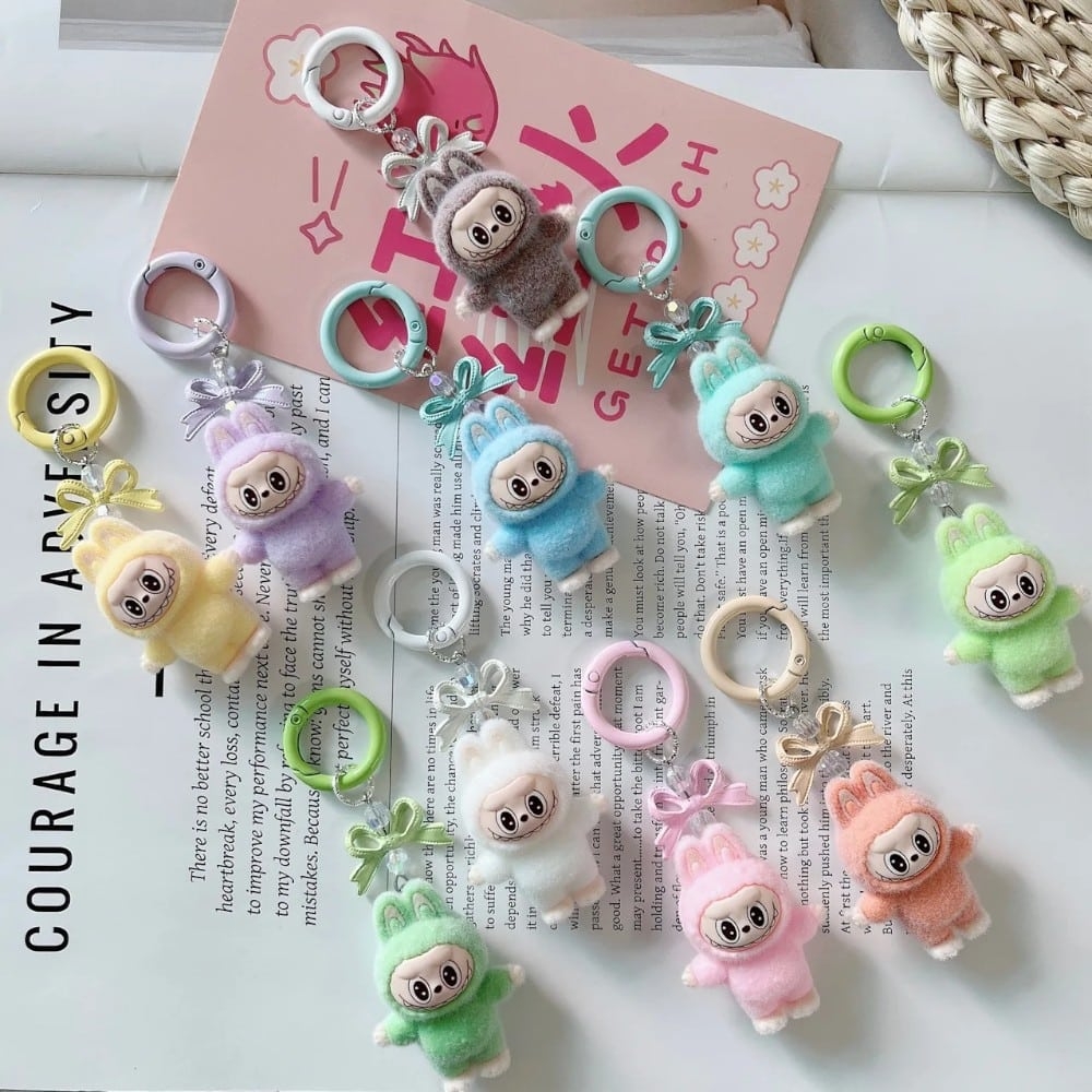 10 mini portachiavi peluche stile Labubu in piedi con anello colorato, in 10 tonalità pastello diverse, disposti su una rivista aperta.