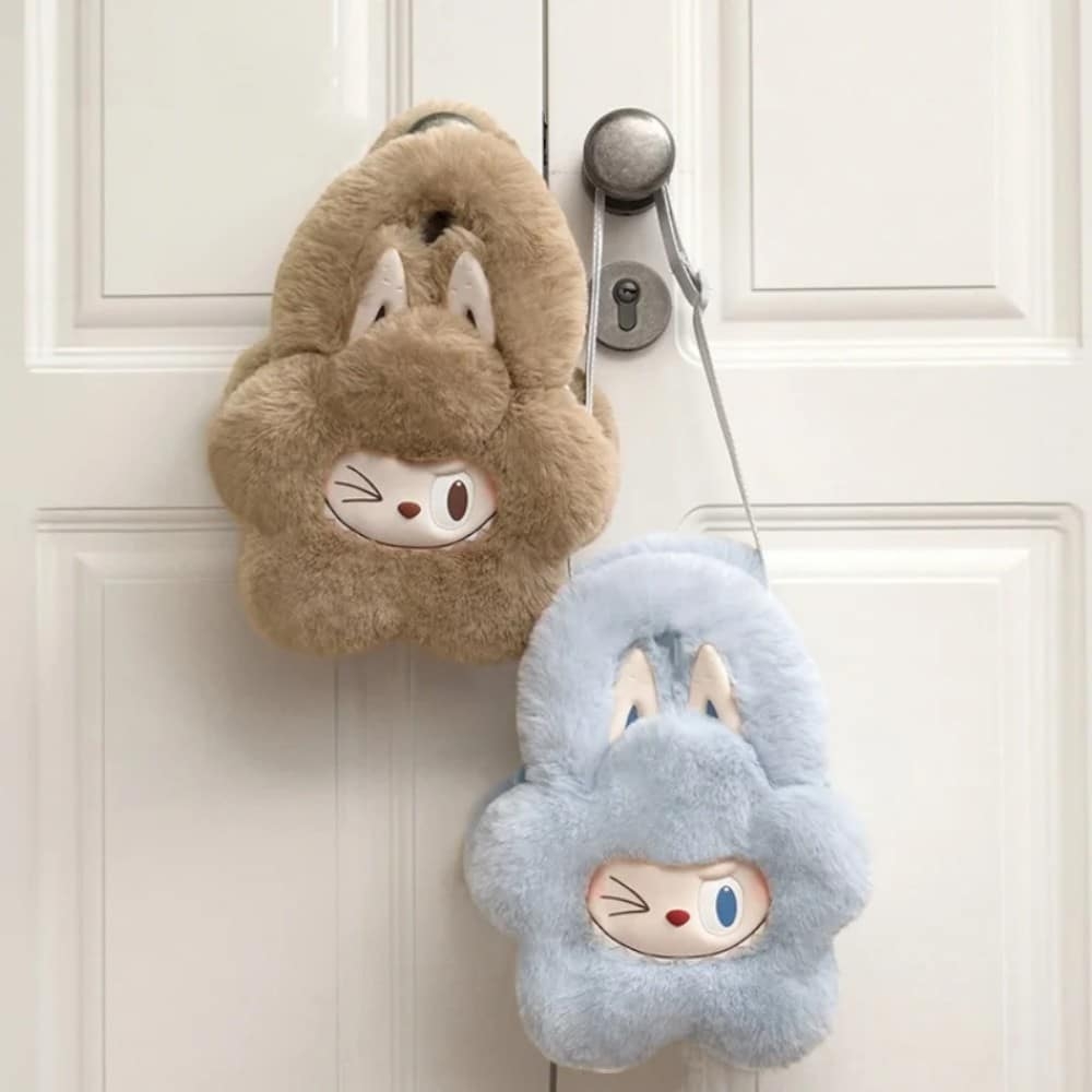 Due piccole borsette morbide a forma di fiore con viso Labubu che strizza l’occhio e grandi orecchie da coniglio; una azzurro cielo e l’altra marrone chiaro. Sono appese a una maniglia.