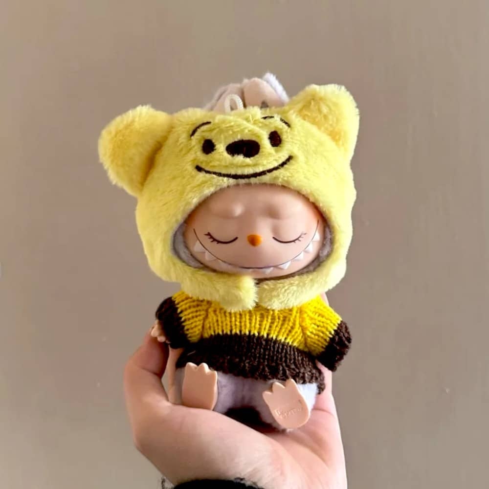 Completo per peluche con cappuccio giallo con orecchie sorridenti e maglione a righe gialle e marroni, indossato da una bambola Labubu addormentata.